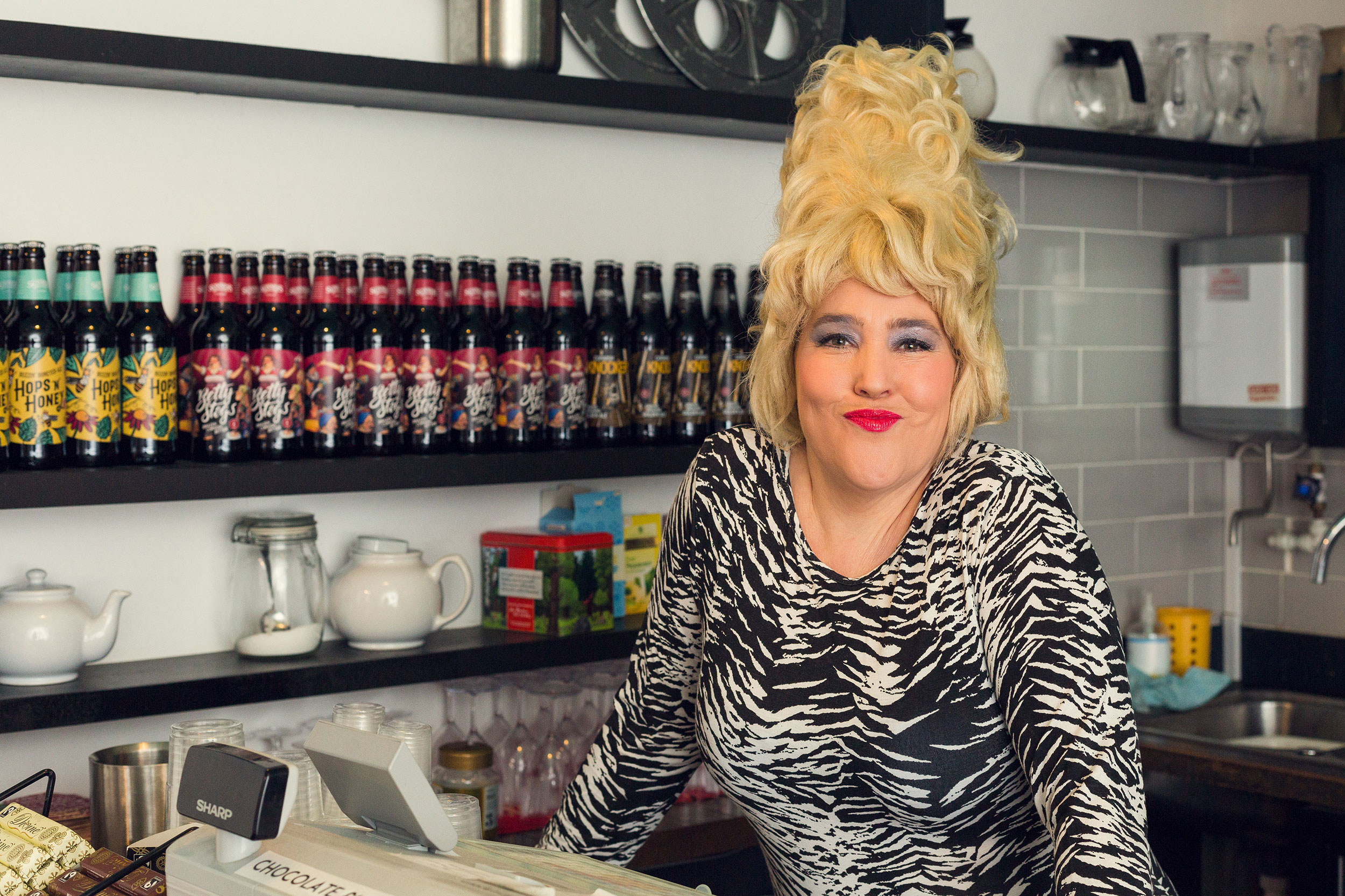 A photograph of a white woman with a tall blonde wig and bright red lipstick standing smiling behind a café bar.