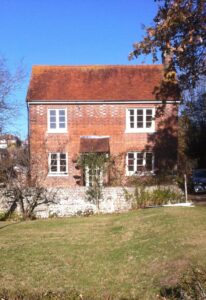 A photograph of Nab Cottage pre-renovation. There is a large green lawn and a white wall in front of the cottage. There is a small roof overhanging the door instead of the new porch. There is bright blue sky and green trees. The cottage is traditional with two windows upstairs two windows downstairs and the doorway between. The tiles of the roof orange and there is a chimney on the right hand side..