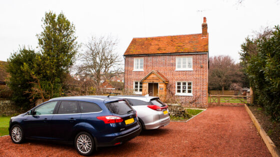 A photograph. At the front are two cars parked on a orange gravel drive in front of a therenovated Nab Cottage, a red brick cottage with an orange tiled roof. To the left and right are mature green trees. The cottage is a traditional style with two windows upstairs above two windows downstairs and a doorway with a porch in the middle. There is a chimney on the right hand side of the roof. To the right of the house is a wooden five bar gate including a personnel gate and a vehicle gate together.