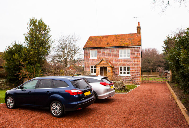 A photograph. At the front are two cars parked on a orange gravel drive in front of a therenovated Nab Cottage, a red brick cottage with an orange tiled roof. To the left and right are mature green trees. The cottage is a traditional style with two windows upstairs above two windows downstairs and a doorway with a porch in the middle. There is a chimney on the right hand side of the roof. To the right of the house is a wooden five bar gate including a personnel gate and a vehicle gate together.